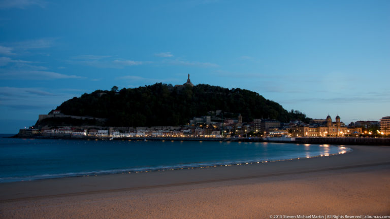 Monte Urgull across calm bay water at blue hour, with shoreline lights and the summit monument visible.