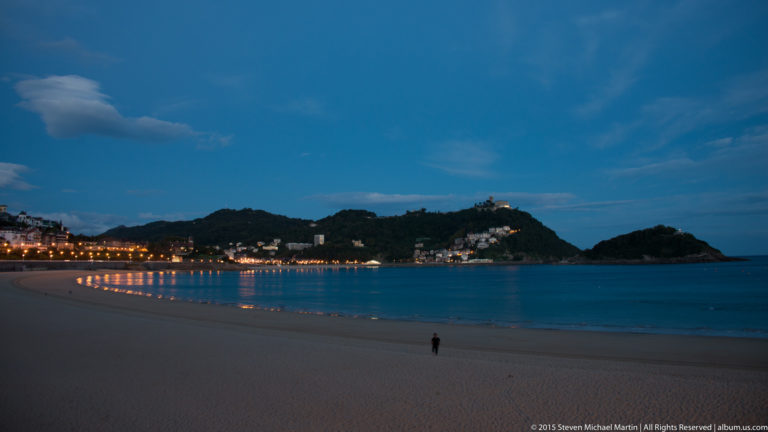 Wide blue-hour view across La Concha Bay, with a curving lit promenade, dark headlands, and one person on the beach.