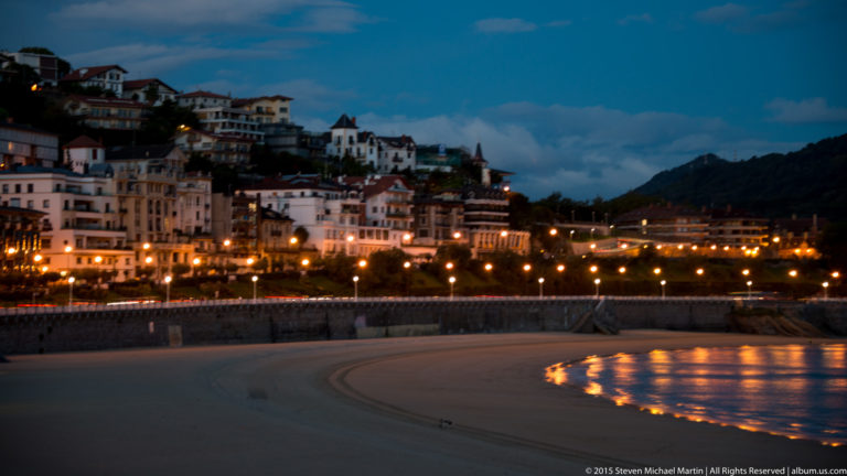 Illuminated waterfront buildings curve around La Concha Bay at dusk, with lights reflected near the sandy shore.