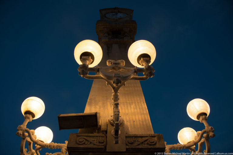 Low-angle close view of ornate glowing globe lamps with a tall promenade clock column behind.