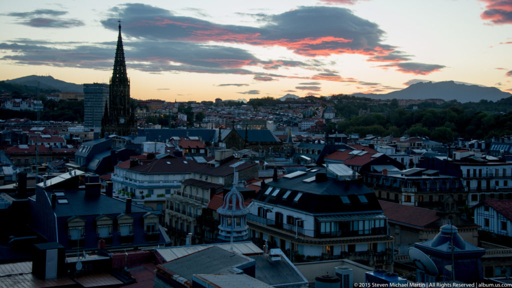 Pointed church spire above dense San Sebastián rooftops beneath a pink-and-blue sunset sky.