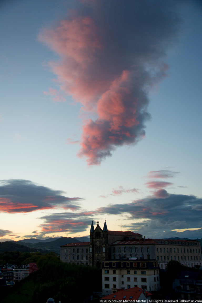 Long dark building with twin pointed chapel towers beneath broad pink sunset clouds.