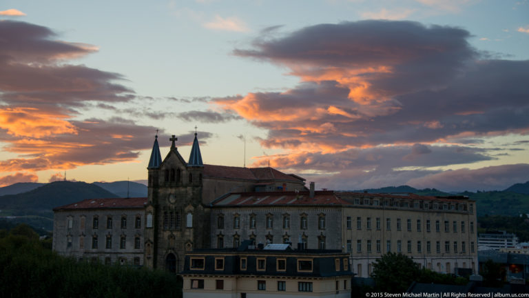 Large historic masonry building with twin pointed chapel towers and crosses beneath orange sunset clouds.