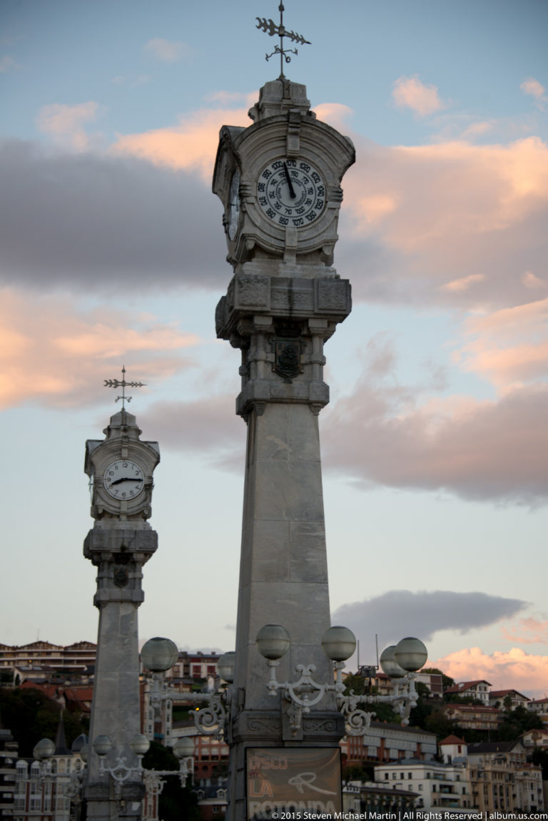 Two tall stone clock columns with weather vanes and globe lamps along the waterfront at sunset.
