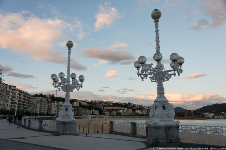 Ornate white multi-globe lamp standards beside the balustrade and beach on La Concha promenade.