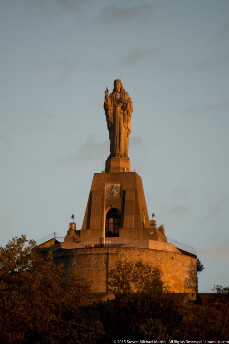Illuminated Sacred Heart statue standing on a high masonry pedestal above Monte Urgull’s fortifications at dusk.