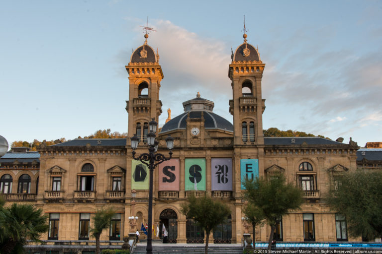 Front view of San Sebastián City Hall with twin towers, central dome, arched windows, banners, and trees.