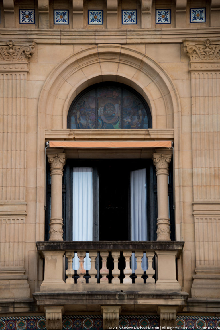 Ornate City Hall window with paired columns, stone balcony balustrade, carved surround, and colorful tile bands.