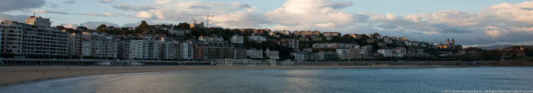 Curving La Concha beachfront lined by multi-story buildings, with a wooded hillside rising behind.