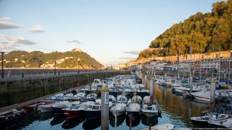 Small boats and sailboats moored in a dense San Sebastián marina, with wooded Monte Urgull rising beyond.