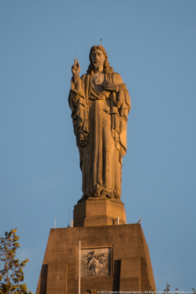 Close view of the robed Sacred Heart statue with raised hand atop its large stone pedestal against blue sky.