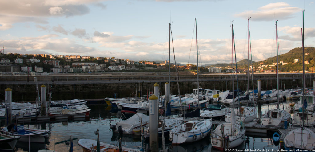 Closely moored sailboats and masts in San Sebastián’s marina, with a built hillside across the harbor.