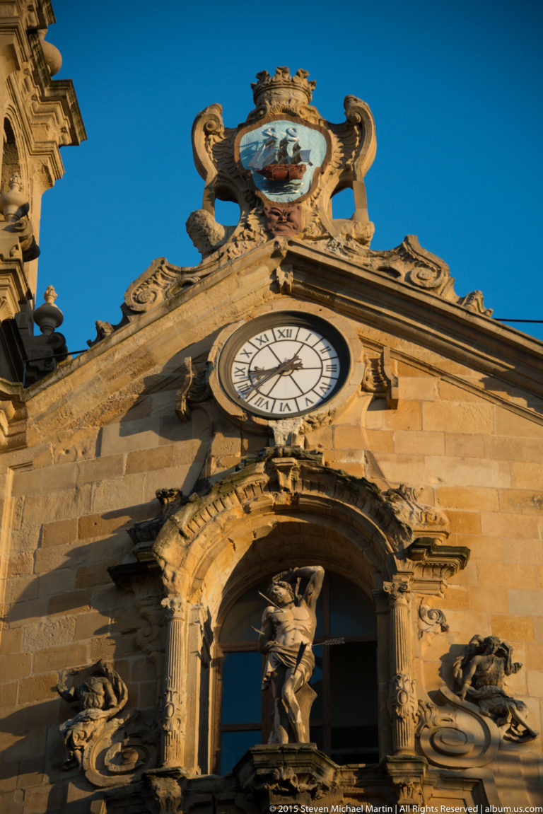 Low-angle stone church façade with Roman-numeral clock, sculpted figure, elaborate carving, and painted ship emblem above.