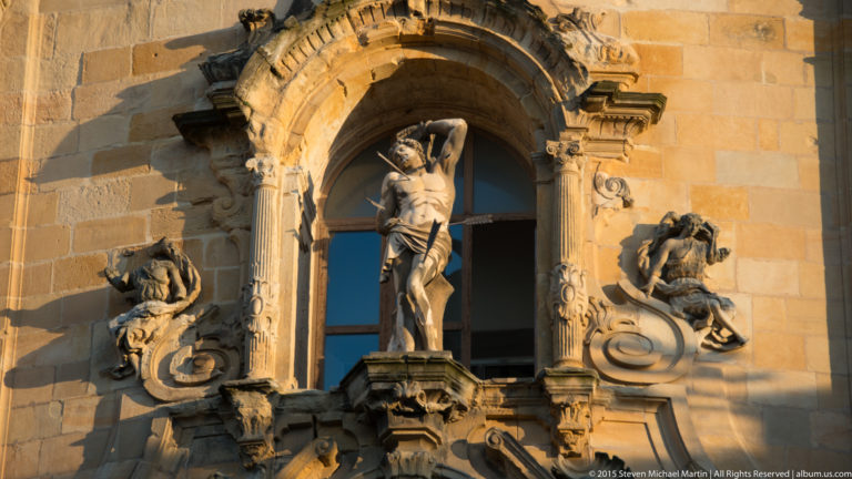 Stone figure standing in an arched niche, surrounded by carved scrolls and smaller figures on an ornate church façade.