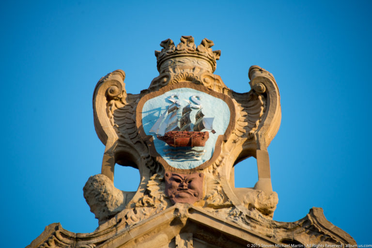 Painted heraldic shield with a sailing ship, framed by carved stone scrolls and a crown-like crest against blue sky.