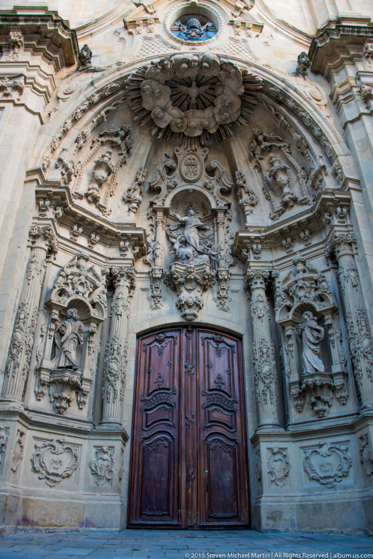 Elaborately carved Baroque stone portal surrounding tall double wooden doors, with niches, statues, reliefs, and scrollwork.