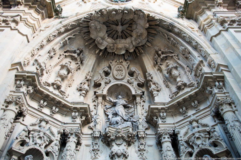 Upper section of an ornate stone church portal with central religious statue, angels, heraldic shields, and radiating carved canopy.