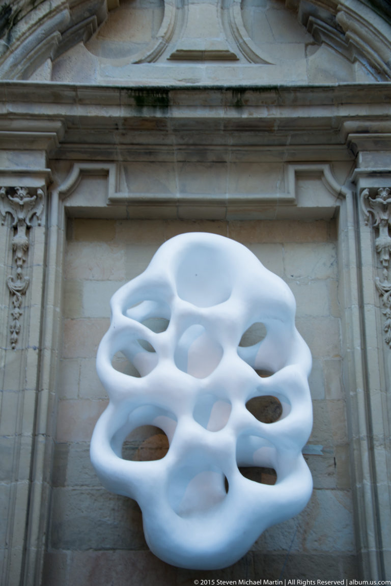 White abstract wall-mounted sculpture with rounded openings set against a pale classical stone façade.