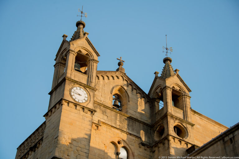 Twin stone bell towers with a round clock, open belfries, bells, and metal finials against blue sky.