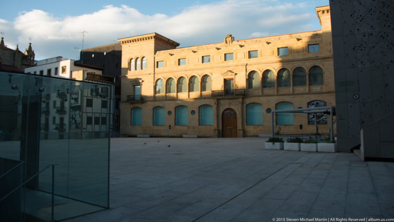 Open paved plaza with a long historic arcaded building, glass structure, and dark modern wall in the foreground.