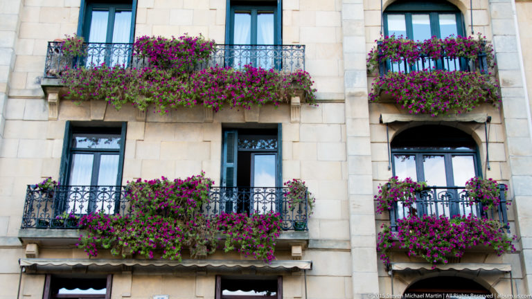 Pale stone façade with four wrought-iron balconies overflowing with bright magenta flowers.