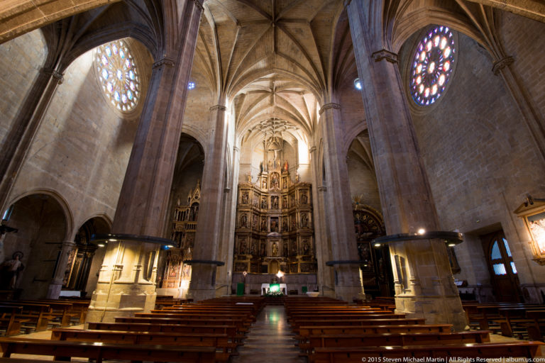 Stone Gothic church nave with pointed vaults, wooden pews, circular stained glass, and a gilded high altar.