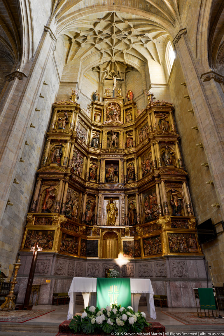Large gilded multi-tiered church retablo filled with framed religious scenes and statues above a white altar.