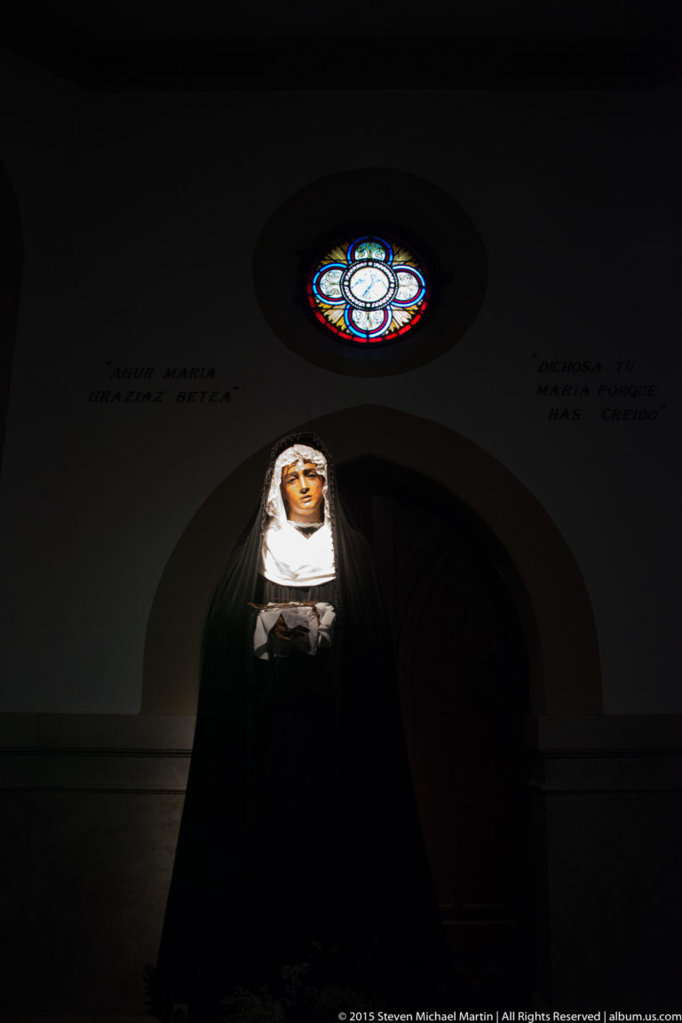 Veiled female religious figure in a dark interior beneath a small circular stained-glass window.