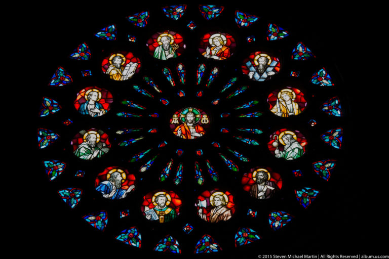 Large circular stained-glass rose window with a central sacred figure surrounded by radiating colored panels and haloed figures.