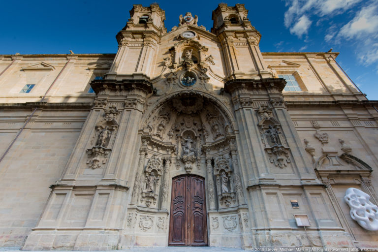 Low-angle view of an ornate stone church façade with wooden doors, dense sculpture, clock, and twin upper structures.