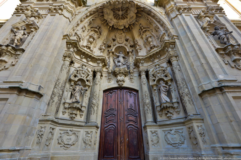 Elaborately carved stone church portal with tall wooden doors, statues, niches, shields, columns, and scrollwork.
