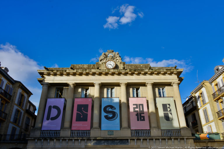 Historic civic building with upper colonnade, central clock and crest, and large colorful vertical banners.