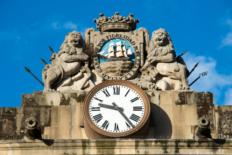 Large Roman-numeral clock beneath a crowned stone coat of arms showing a sailing ship between two sculpted lions.
