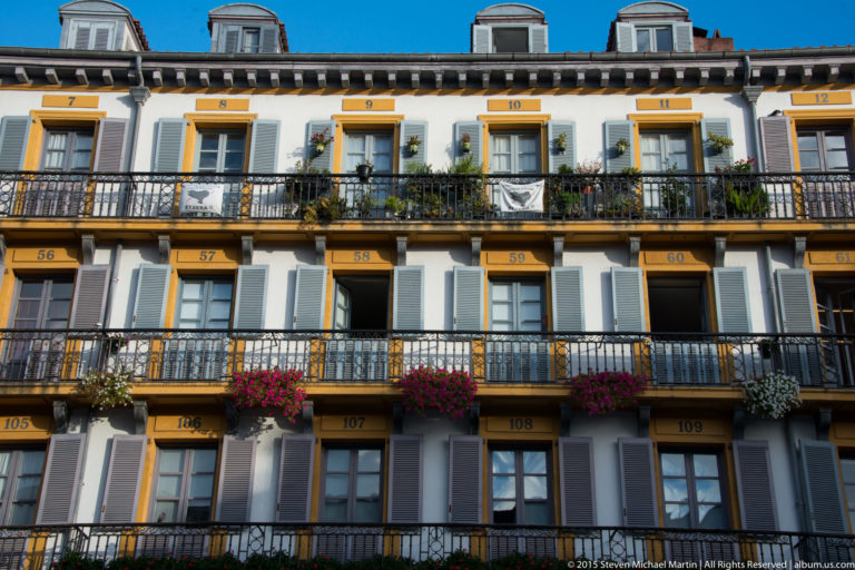 Multi-story pale residential façade with numbered window bays, gray shutters, continuous iron balconies, and flower boxes.