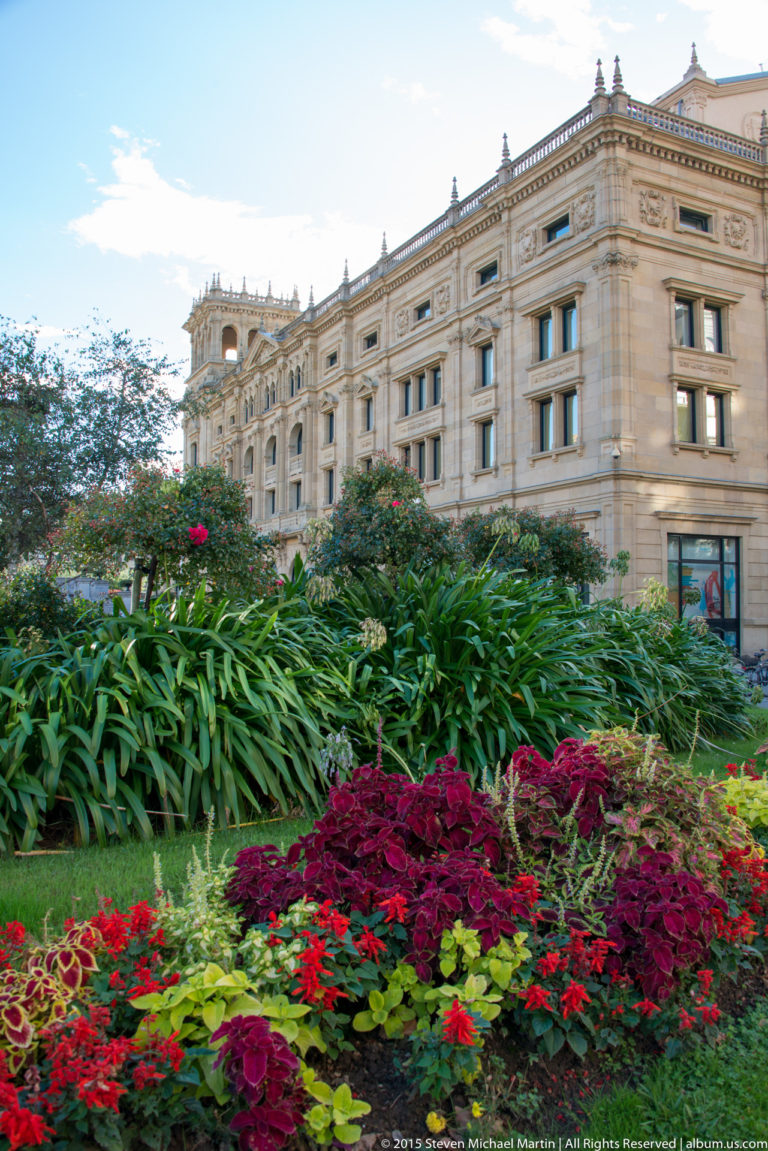 Colorful formal flower beds and tall foliage in front of a large ornate pale-stone civic building.