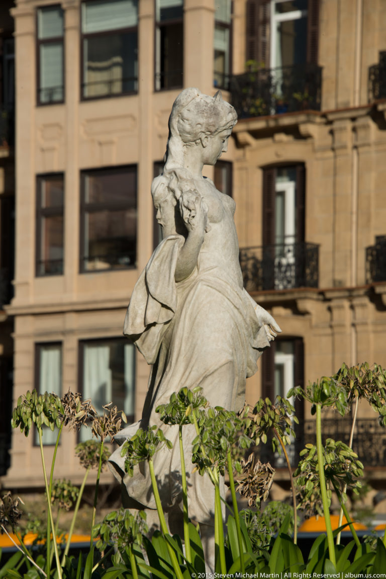 Pale stone sculpture of a draped woman seen among tall green plants, with an apartment façade behind.