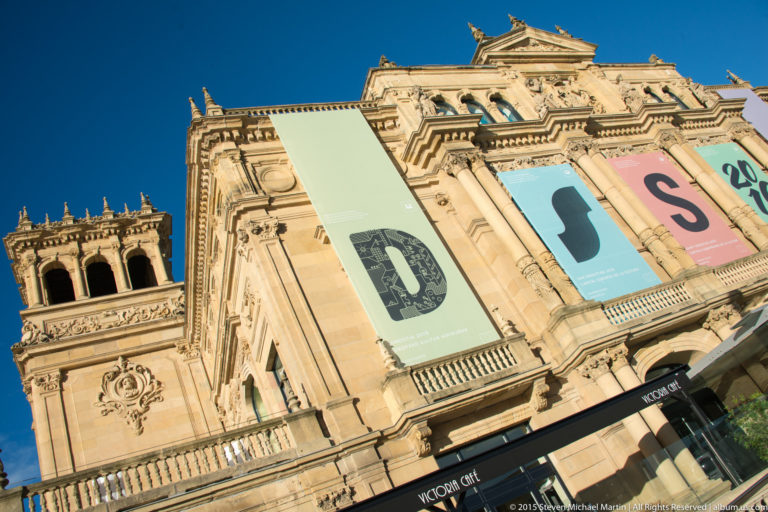 Low-angle view of an ornate pale-stone cultural building with columns, tower, pediments, and large vertical exhibition banners.