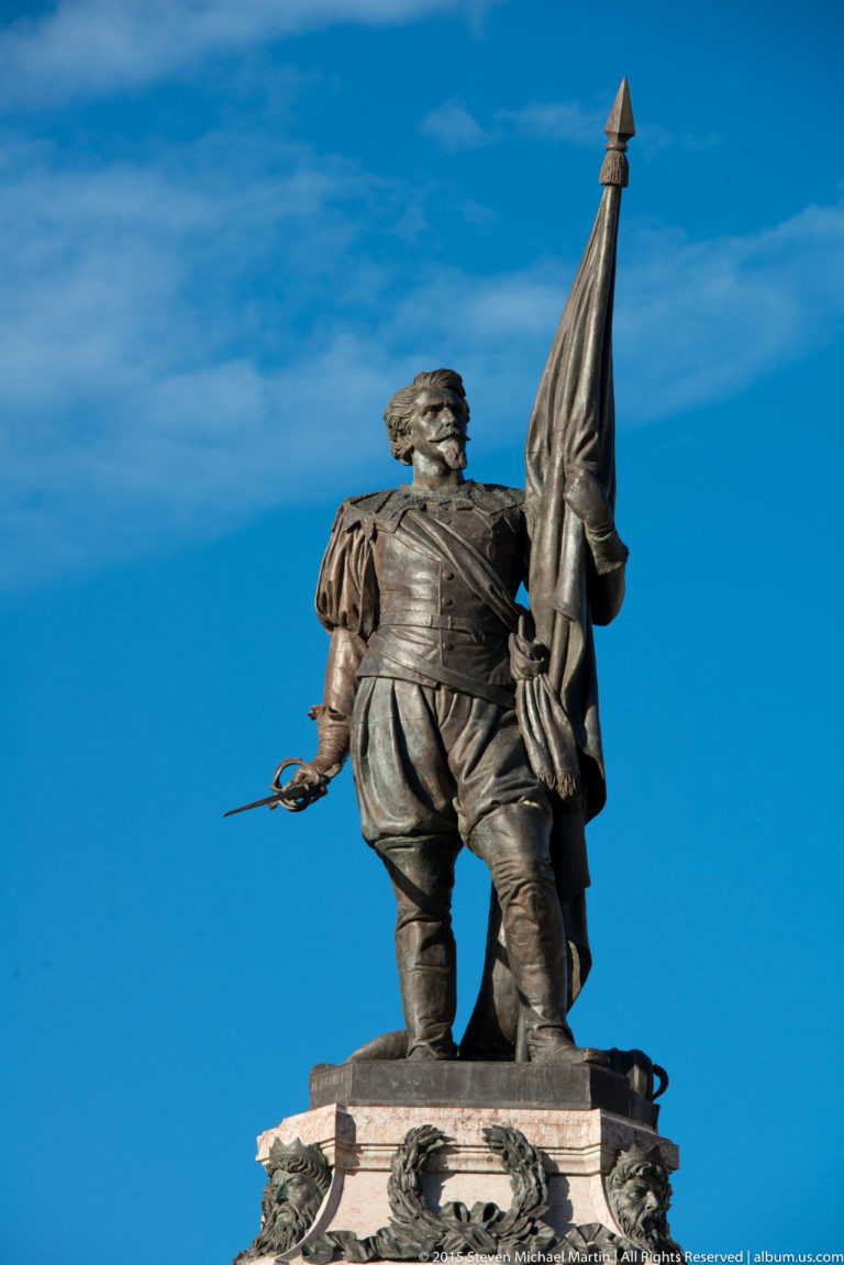 Low-angle bronze statue of a historical male figure holding a large standard on a stone pedestal against blue sky.