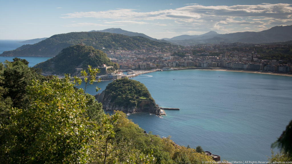 Elevated panorama over San Sebastián’s blue bay, wooded Monte Urgull, dense waterfront city, and mountains beyond.