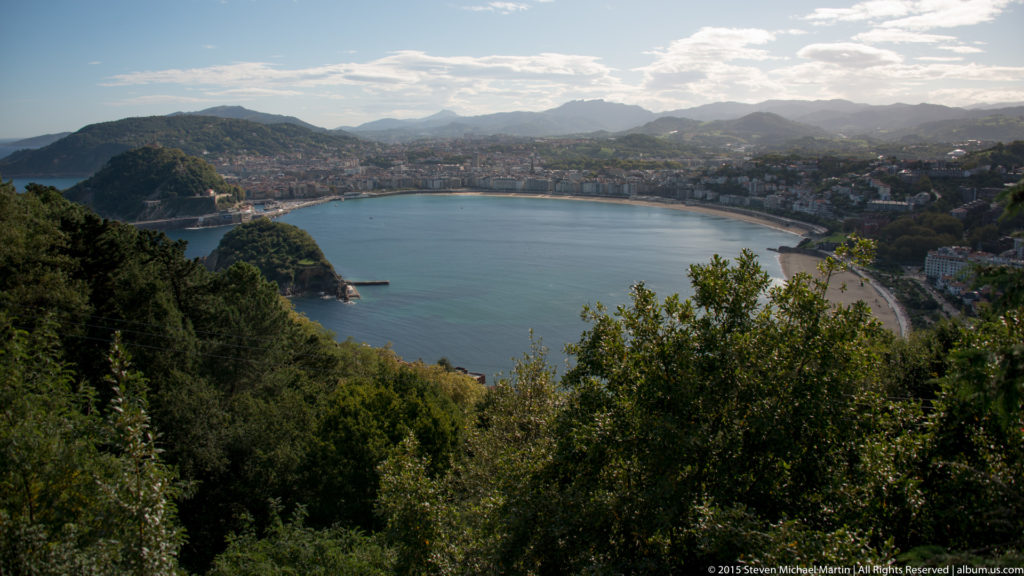 High view across La Concha Bay, framed by trees, with curving beaches, wooded headlands, city buildings, and distant mountains.
