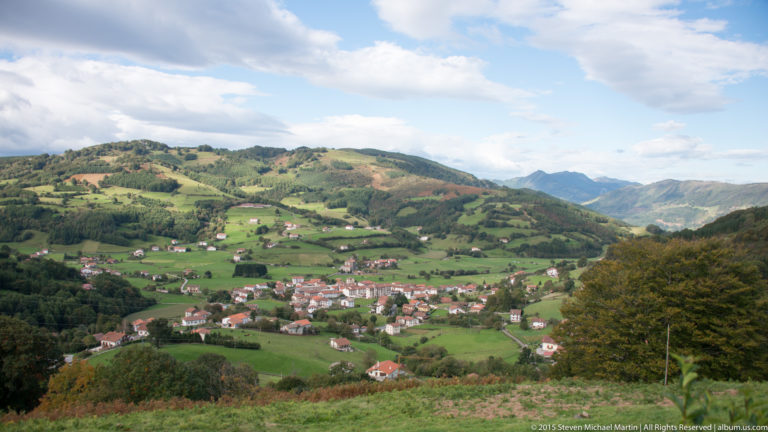 Green Basque valley with a compact red-roofed village, scattered farmhouses, fields, wooded slopes, and distant mountains.