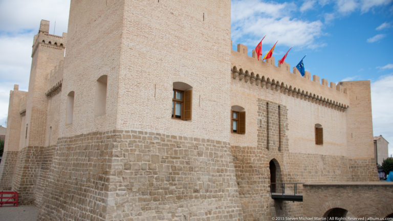 Restored pale-stone fortress with square towers, crenellated parapet, flags, and a bridge leading to an arched entrance.