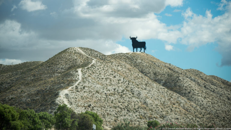 Large black bull silhouette standing on a pale barren ridge beneath blue sky and heavy clouds.