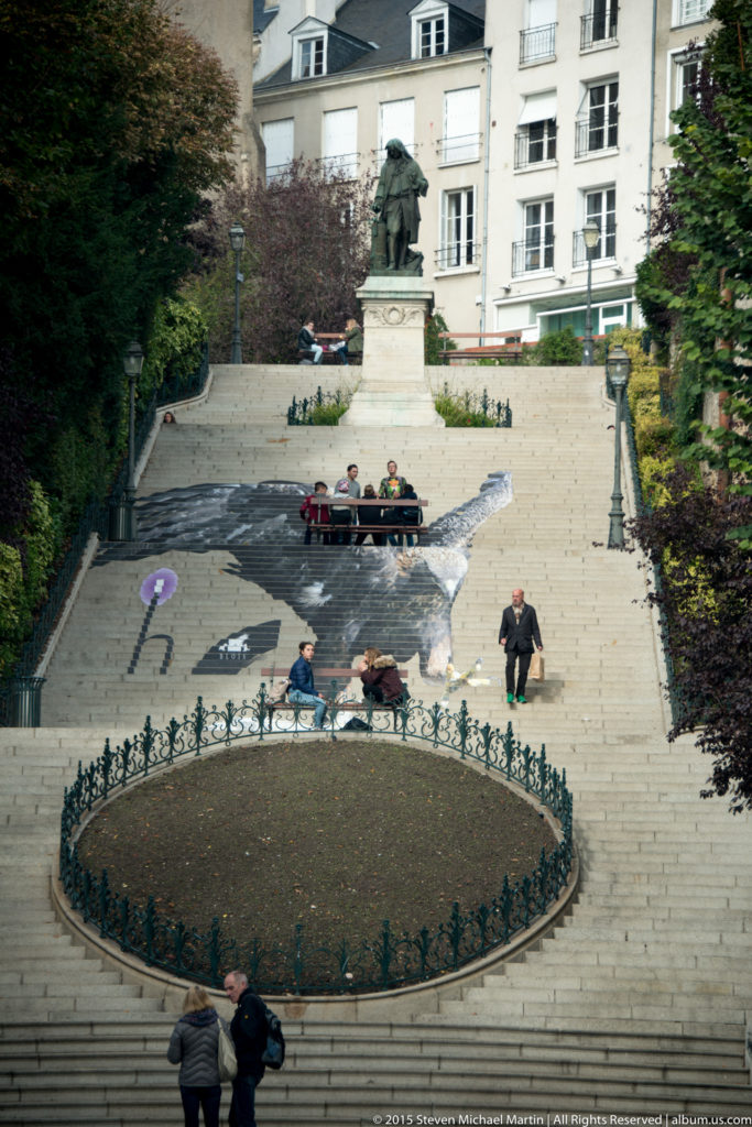 Elevated view of a formal square with monument, stone steps, planting beds, and townhouses in Blois.
