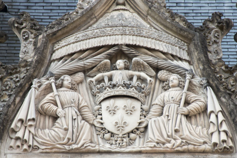 Carved crowned fleur-de-lis shield flanked by angelic figures beneath an ornate architectural canopy.