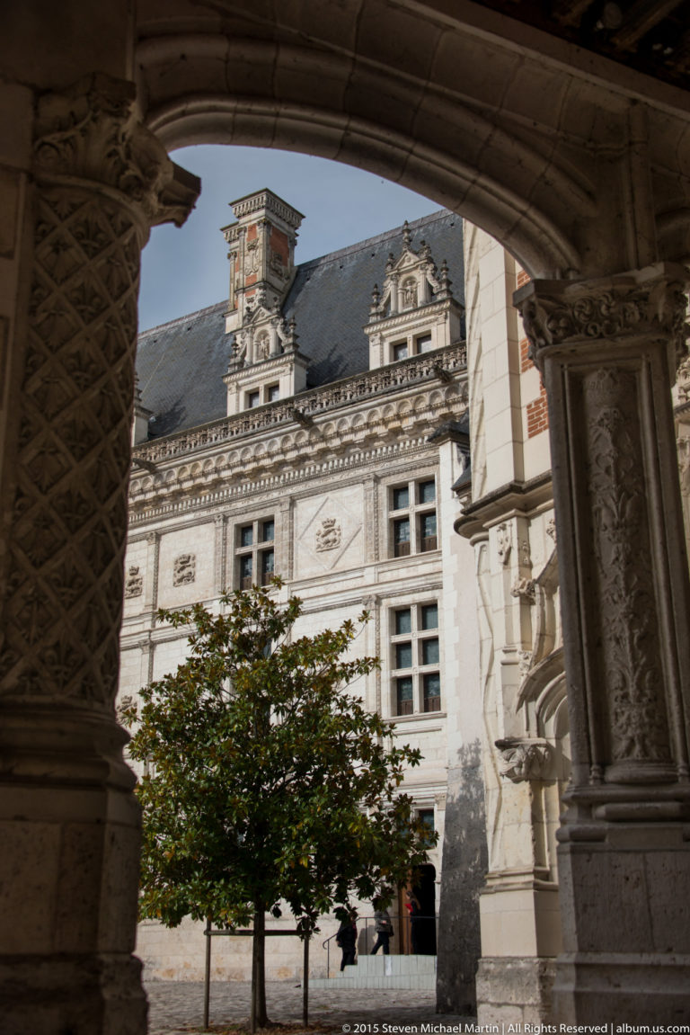 Sunlit Renaissance château façade and courtyard seen through a dark stone arcade in Blois.