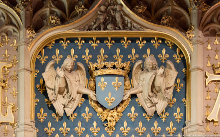 Crowned blue fleur-de-lis shield held by two sculpted angels within ornate gilded interior decoration.