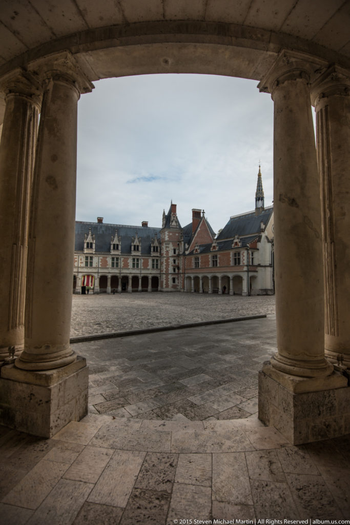 Blois château courtyard viewed through large stone columns, with brick façades and a spire beyond.