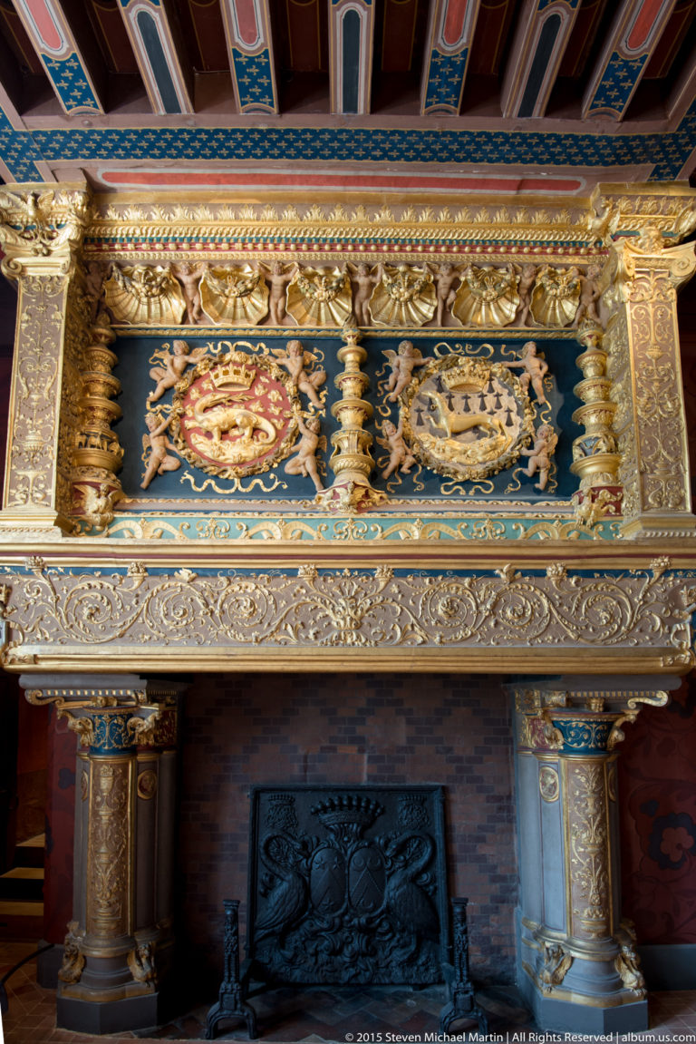 Ornate château fireplace with gilded carving, heraldic panels, polychrome decoration, and dark iron fireback.