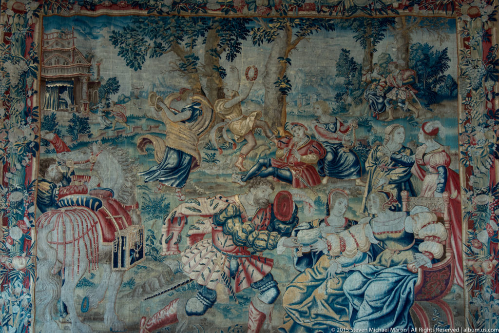 Large historic tapestry showing many figures, horses, architecture, and landscape within a decorative border.